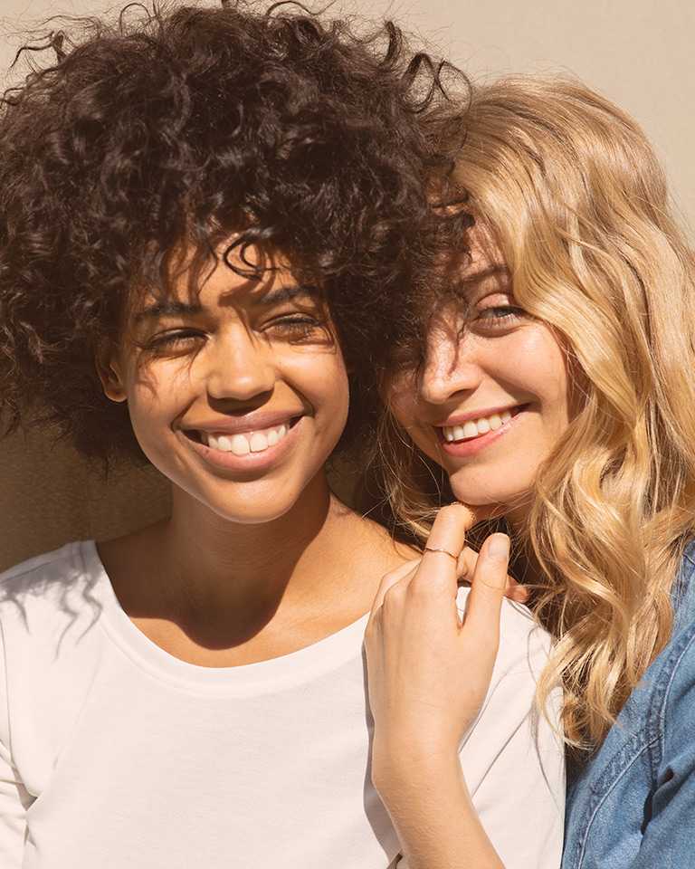 Female with dark curly hair and female with blonde wavy hair laughing in the sun 